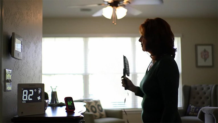Homeowner checking thermostat set to cool while room reads 82°F, using a hand fan in summer light.