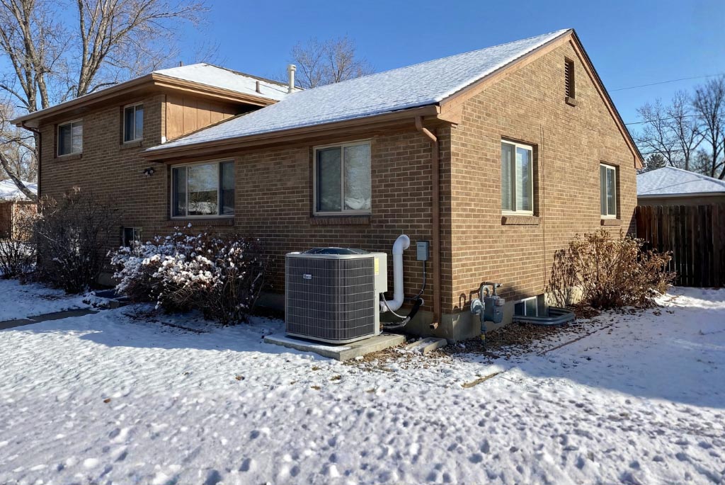 Brick Denver home in winter with light snow and a modern outdoor heat pump unit and gas meter on the side wall, illustrating a dual-fuel heating setup.