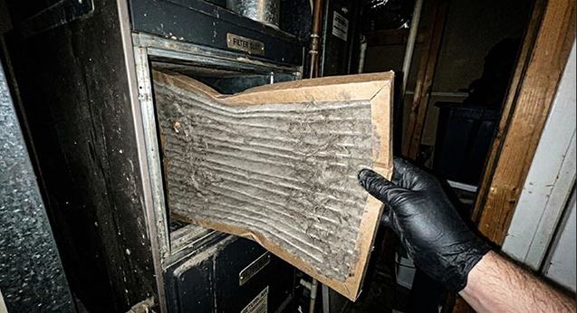 A first-person view of a hand in a black nitrile glove pulling a severely dirty, dust-caked, and physically bent pleated air filter out of a furnace filter slot in a dimly lit utility closet.