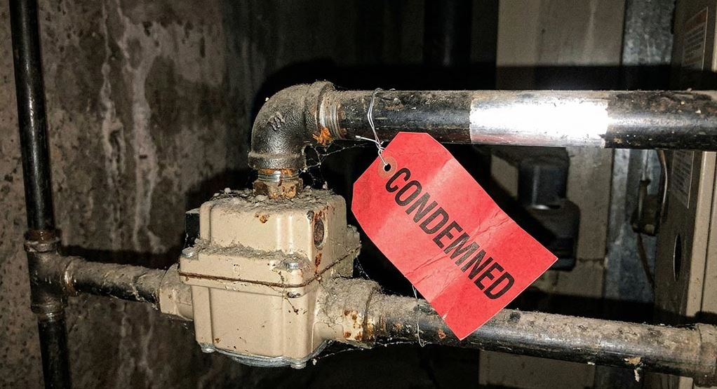 A close-up view of grimy, corroded furnace piping and a gas valve in a dark basement setting. A prominent bright red inspection tag with the word 'CONDEMNED' is wire-tied to the pipe.