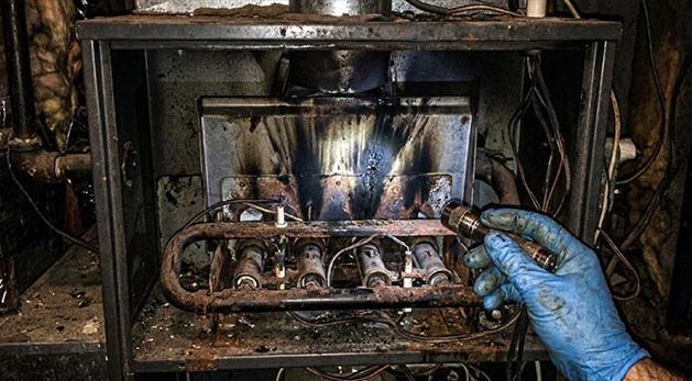 A close-up photograph shows a technician's hand, in a blue glove, holding a flashlight to closely inspect the rusted and heavily soot-stained burner assembly of a condemned furnace.