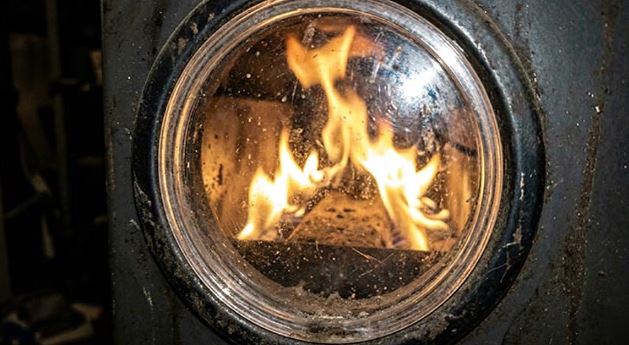 A close-up macro shot looking through a dirty circular sight glass on a metal furnace door, showing dangerous, lazy yellow and orange flames burning inside the chamber instead of safe blue flames.